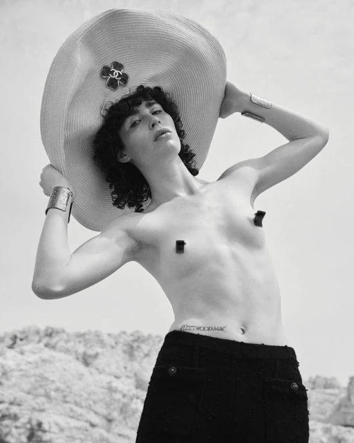 Person poses in a black skirt with a large straw hat on a bright, sunny day. The individual is standing outside near a rocky hill.