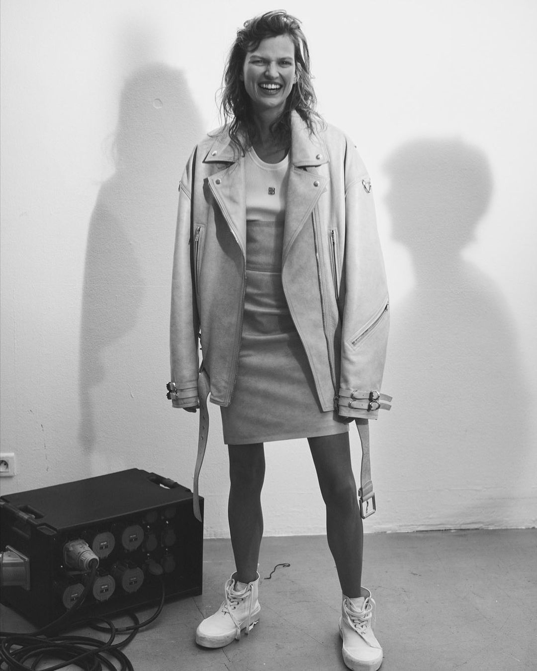 Black and white shot of smiling woman in oversized leather jacket, mini dress, and boots in a minimalist interior setting.