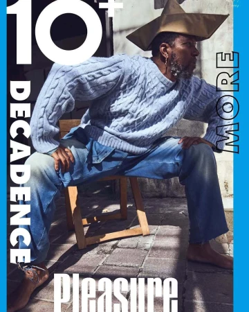 Man wearing a light blue cable knit sweater and denim jeans seated on a wooden chair outdoors, wearing a distinctive geometric hat.