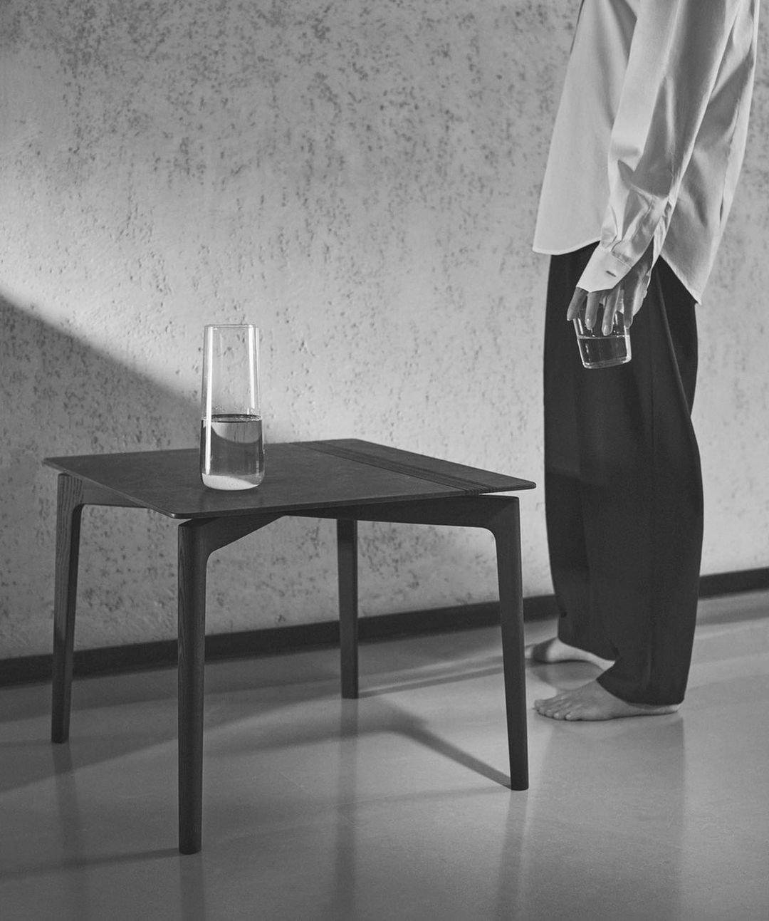 Monochrome portrait of a person standing beside a modern dark table holding a glass of water, photographed by Stefano Galuzzi.