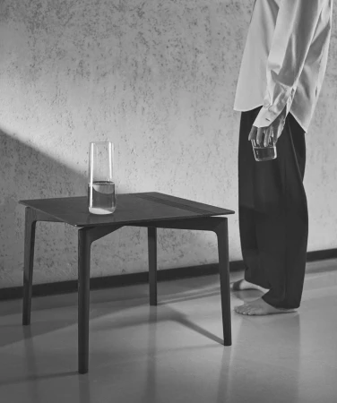Monochrome portrait of a person standing beside a modern dark table holding a glass of water, photographed by Stefano Galuzzi.