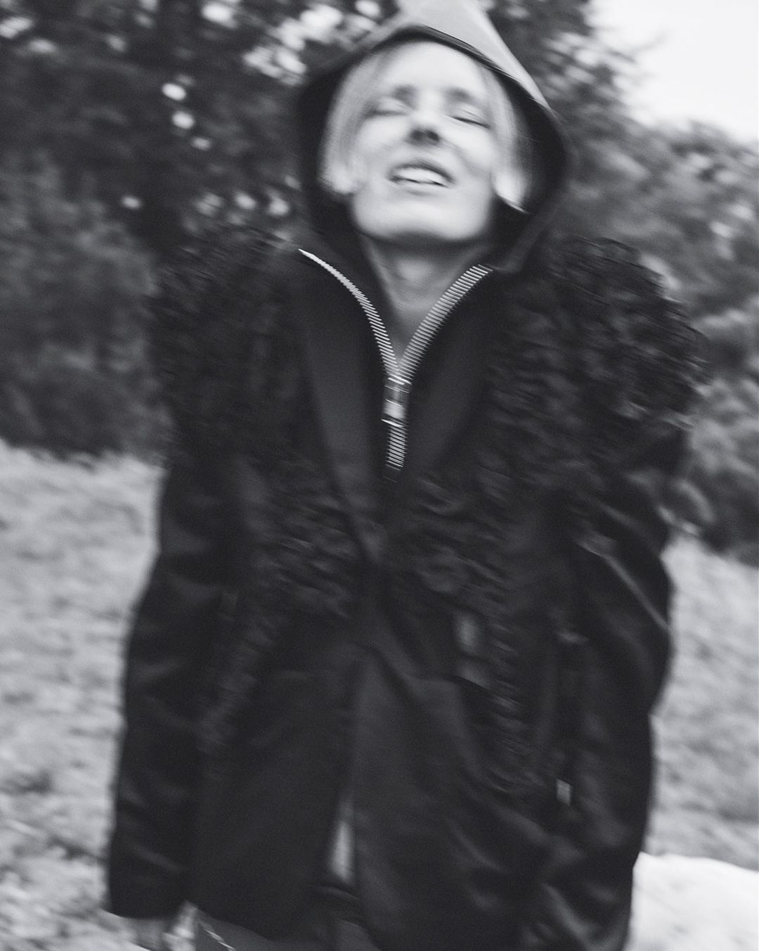 Monochrome portrait of a person with blond hair wearing a hooded fur-trimmed jacket standing outside with trees in the background.