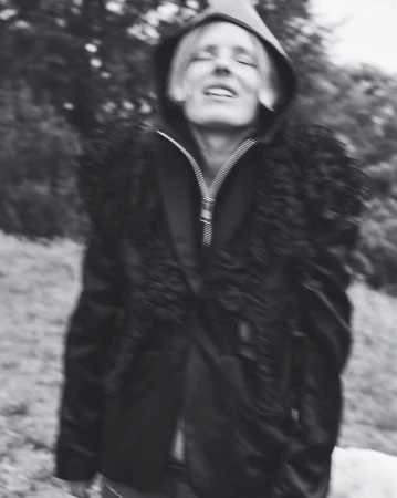 Monochrome portrait of a person with blond hair wearing a hooded fur-trimmed jacket standing outside with trees in the background.