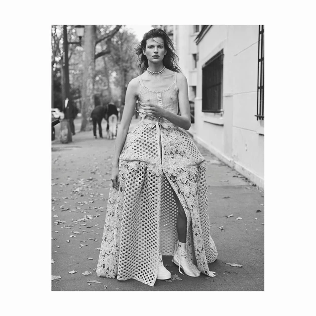 A woman stands on a city street, wearing a buttoned tank top, pearl choker, lace skirt, and white boots, with people and buildings in the background.