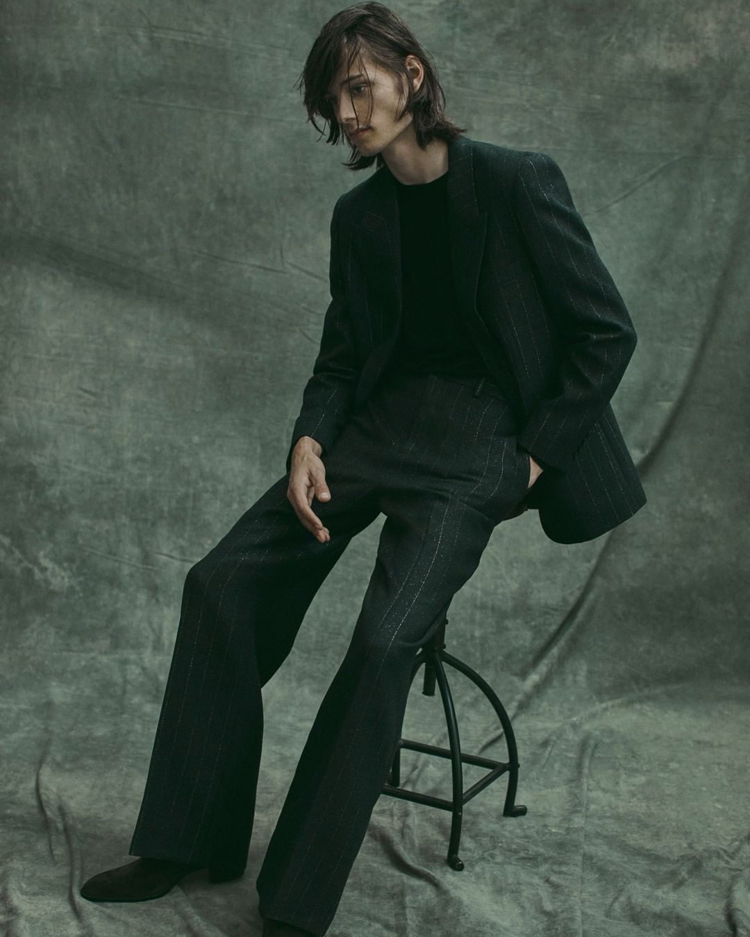 A slender adult male sits on a stool in a pinstripe suit with long hair, posing in a studio against a mottled green backdrop.