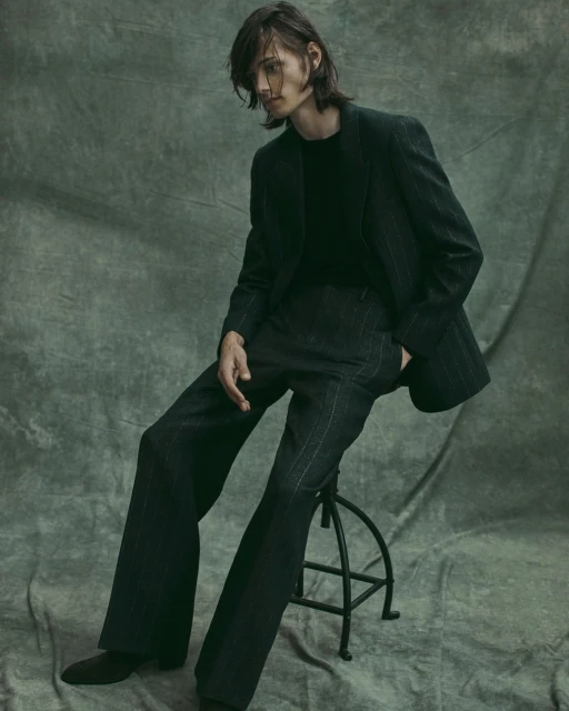 A slender adult male sits on a stool in a pinstripe suit with long hair, posing in a studio against a mottled green backdrop. 