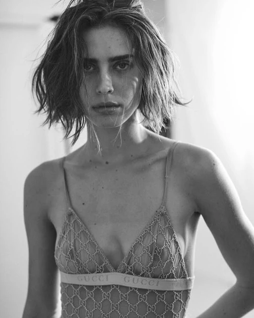 Black and white portrait of a young adult female model in a lace lingerie top with a Gucci logo against a blurred light background.