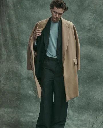 A man wearing a camel overcoat draped over his shoulders, dark trousers, and a blue t-shirt against a textured grey green backdrop.