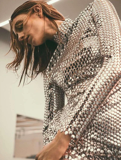 A fashionable adult female poses indoors wearing a shiny, textured, metallic silver long-sleeve dress against a blurred neutral background.