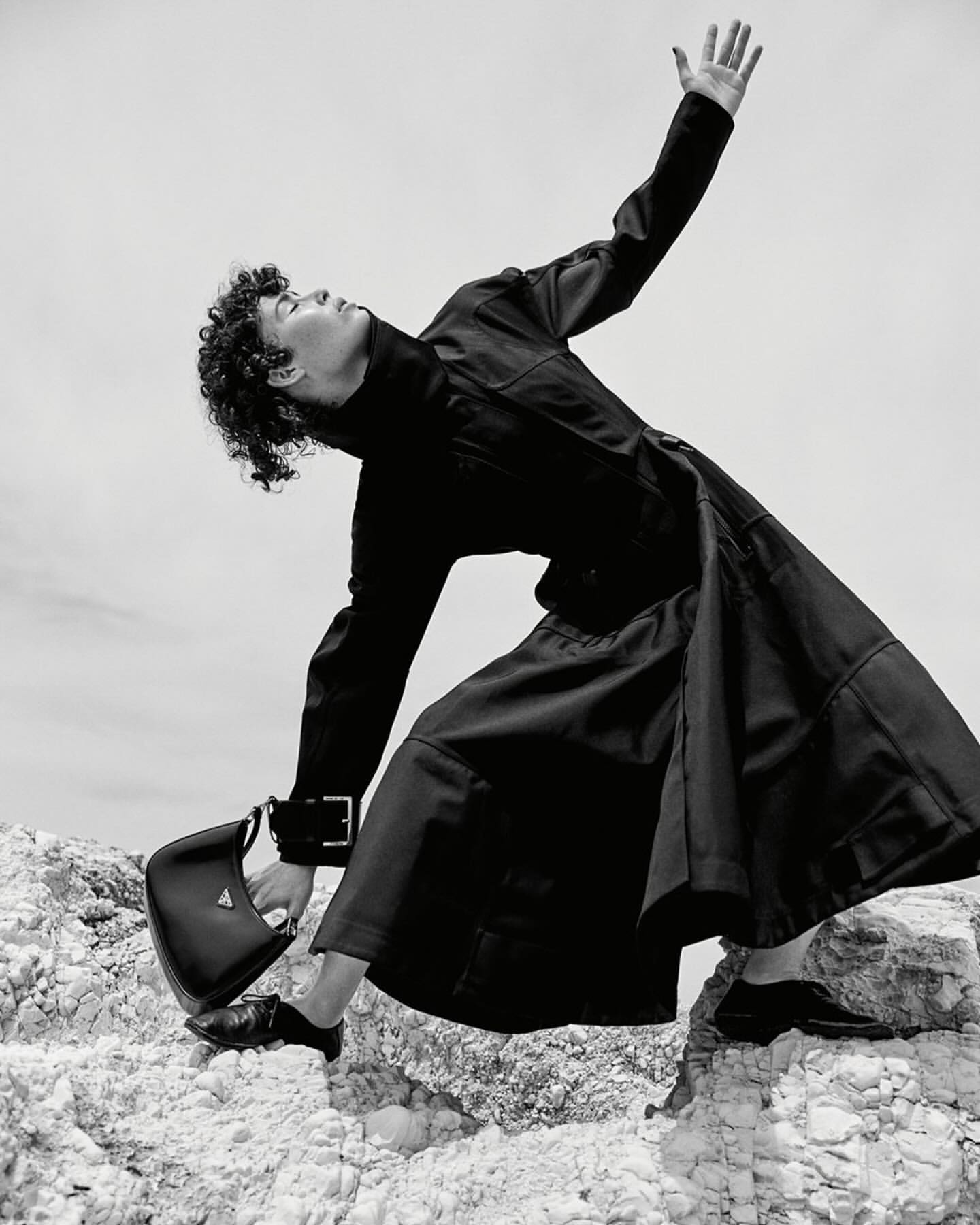 Monochrome fashion shot of person in black coat and dress on rocky terrain, holding Prada purse, against a light sky backdrop.