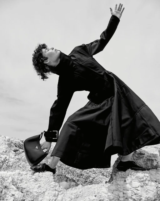 Monochrome fashion shot of person in black coat and dress on rocky terrain, holding Prada purse, against a light sky backdrop.