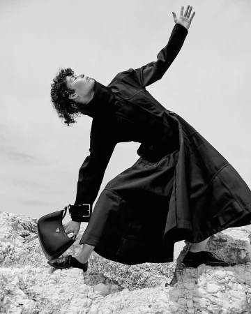 Monochrome fashion shot of person in black coat and dress on rocky terrain, holding Prada purse, against a light sky backdrop.