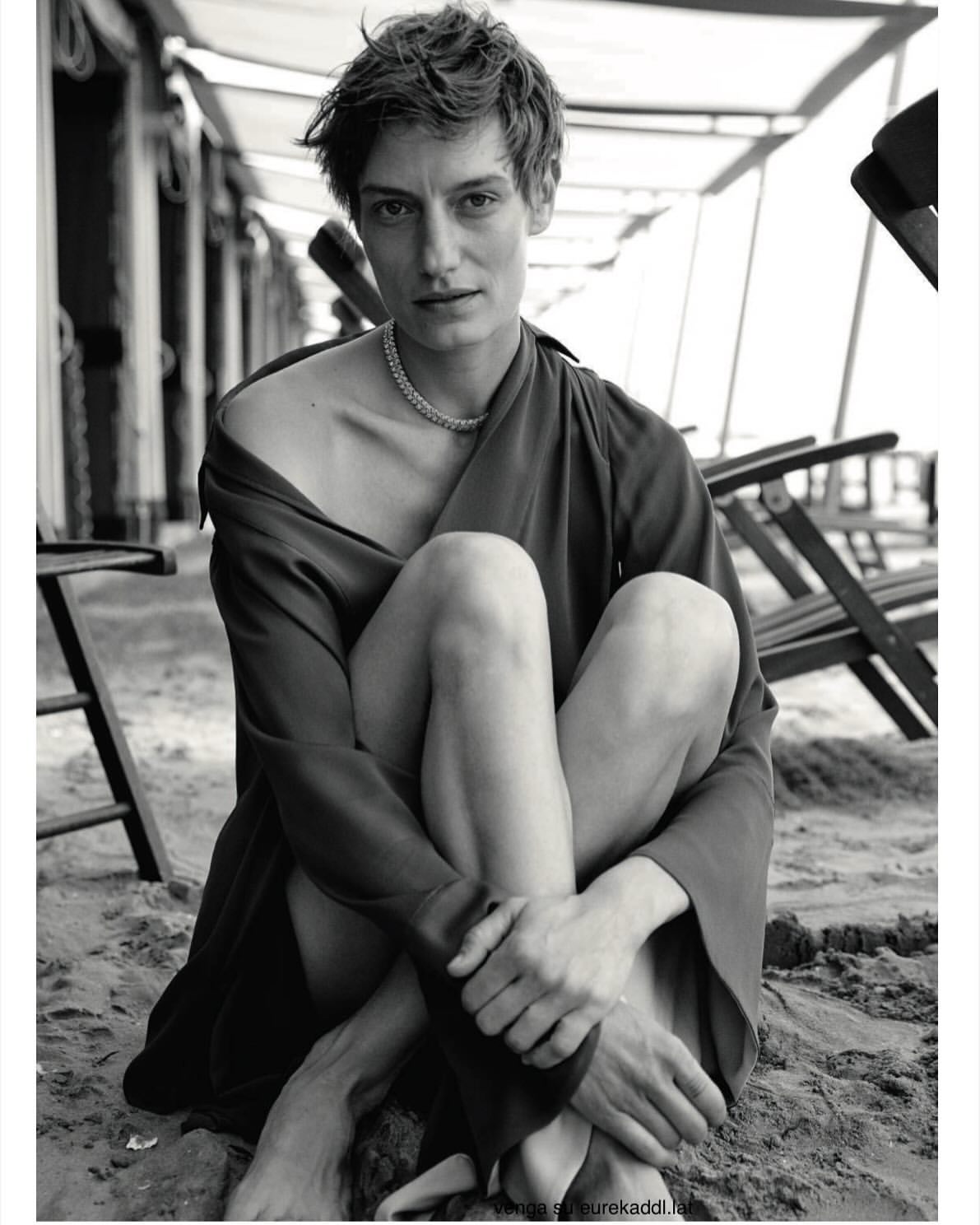 Model with short hair wearing a dark draped dress and necklace seated on sand outdoors by beach chairs.