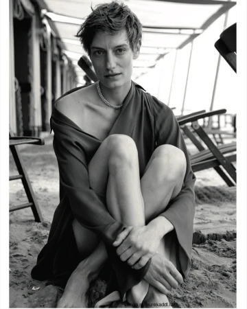 Model with short hair wearing a dark draped dress and necklace seated on sand outdoors by beach chairs.