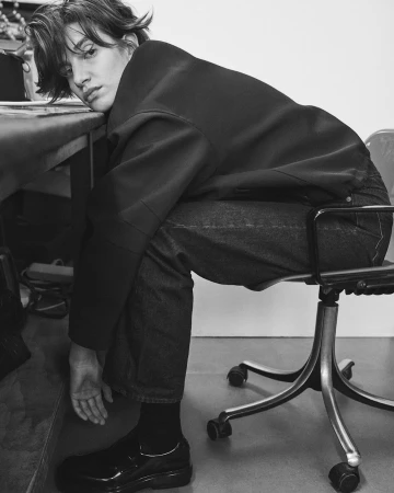 Black and white portrait of a woman resting her head on a table while seated in an office chair.