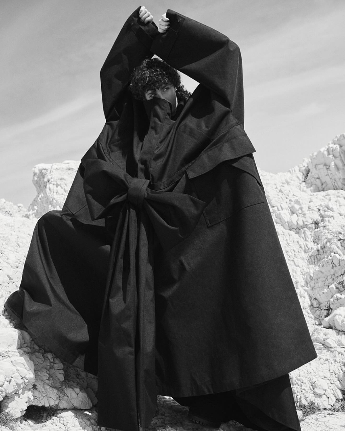 Model in oversized black hooded coat with large front bow standing among rocky terrain, photographed by Stefano Galuzzi