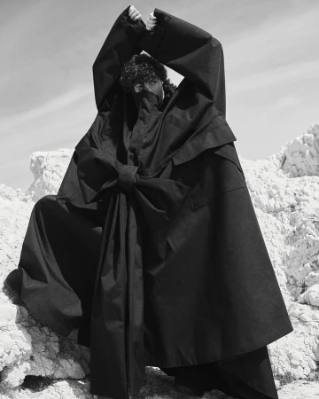 Model in oversized black hooded coat with large front bow standing among rocky terrain, photographed by Stefano Galuzzi