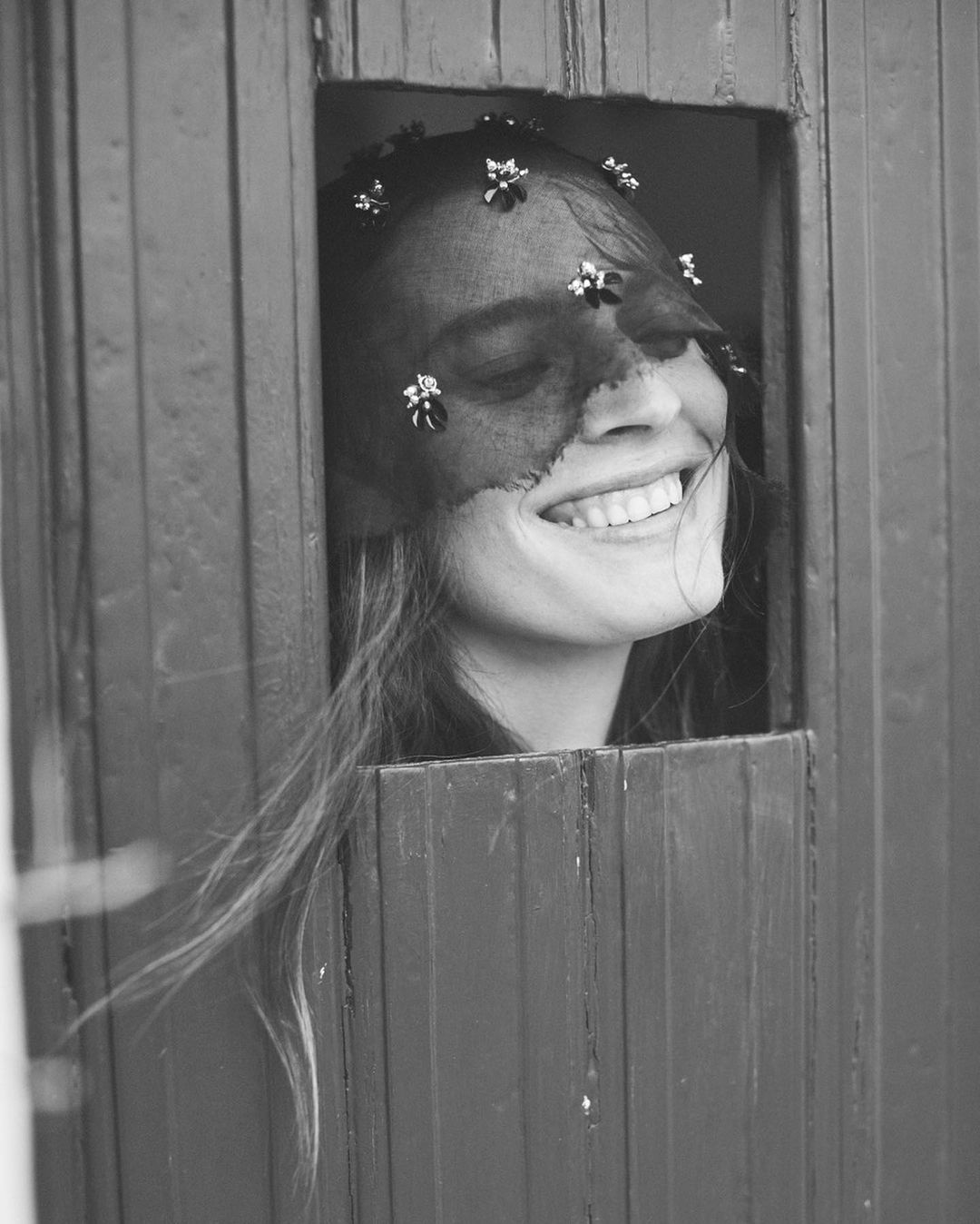 Close-up black-and-white portrait of a smiling woman with long hair, wearing a floral-accented veil and framed by a doorway or window.