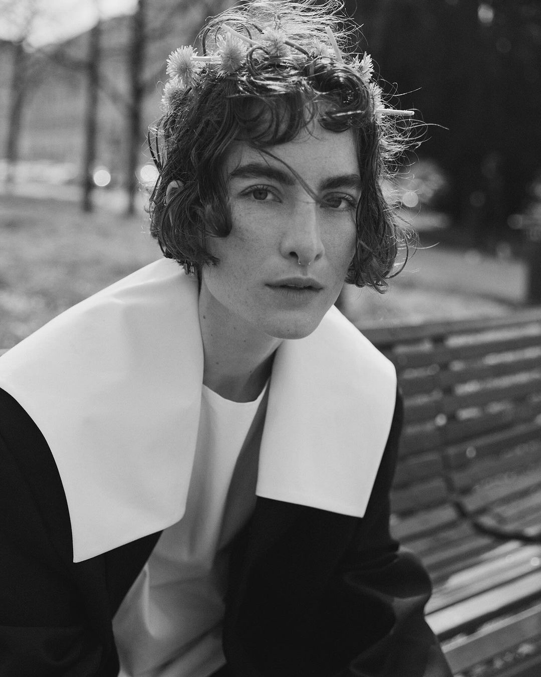 Black and white portrait of a woman wearing a floral crown, wide-collared shirt, and jacket. Shot outdoors on a bench in soft, natural light.