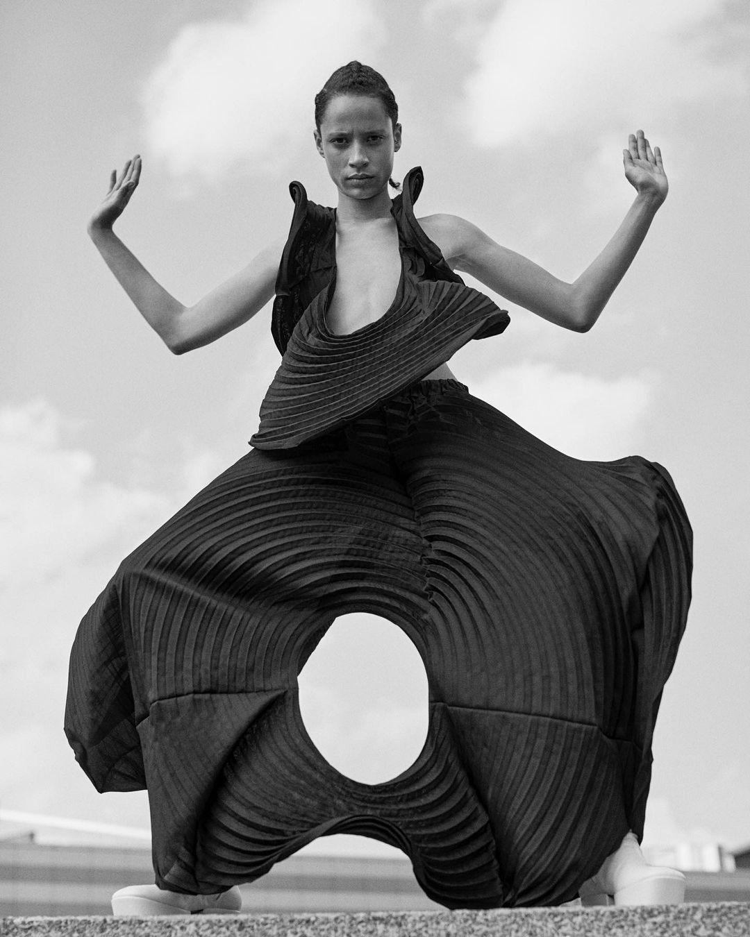 A black-and-white fashion shot features a slender adult with raised hands wearing a dark, geometrically sculpted outfit with large circular openings.
