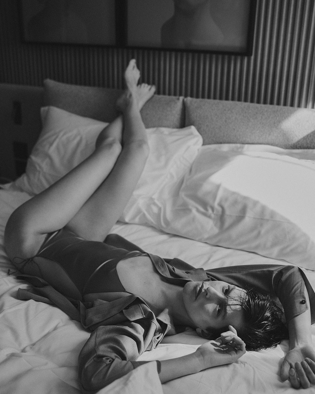 Black and white portrait of a woman lying on a bed, legs raised, wearing a button-down shirt, with soft lighting in an indoor setting.