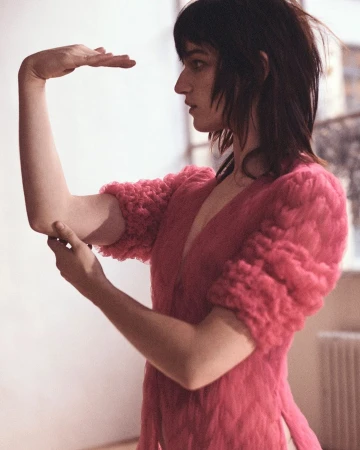 Woman in pink knit sweater poses with raised hands in an indoor photo studio with natural lighting and a cream background and radiator.