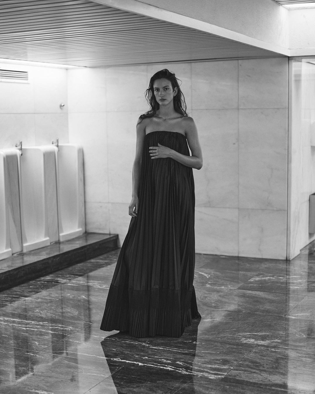 Elegant woman poses in a floor-length black strapless dress in an indoor marble setting with a minimalist architectural backdrop.