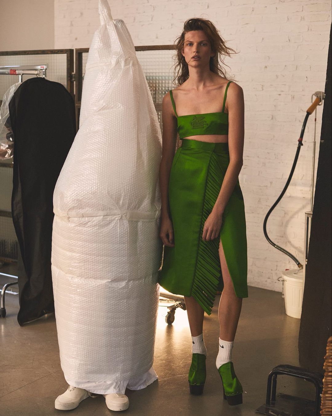 Woman posing in green two-piece skirt set in studio, standing next to large bubble-wrapped object, wearing green platform shoes and white socks.