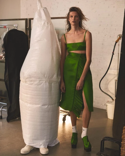 Woman posing in green two-piece skirt set in studio, standing next to large bubble-wrapped object, wearing green platform shoes and white socks.