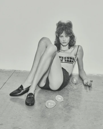 Young adult woman sits on a floor wearing a patterned bra top, shorts, and loafers next to two white balloons in a studio setting.