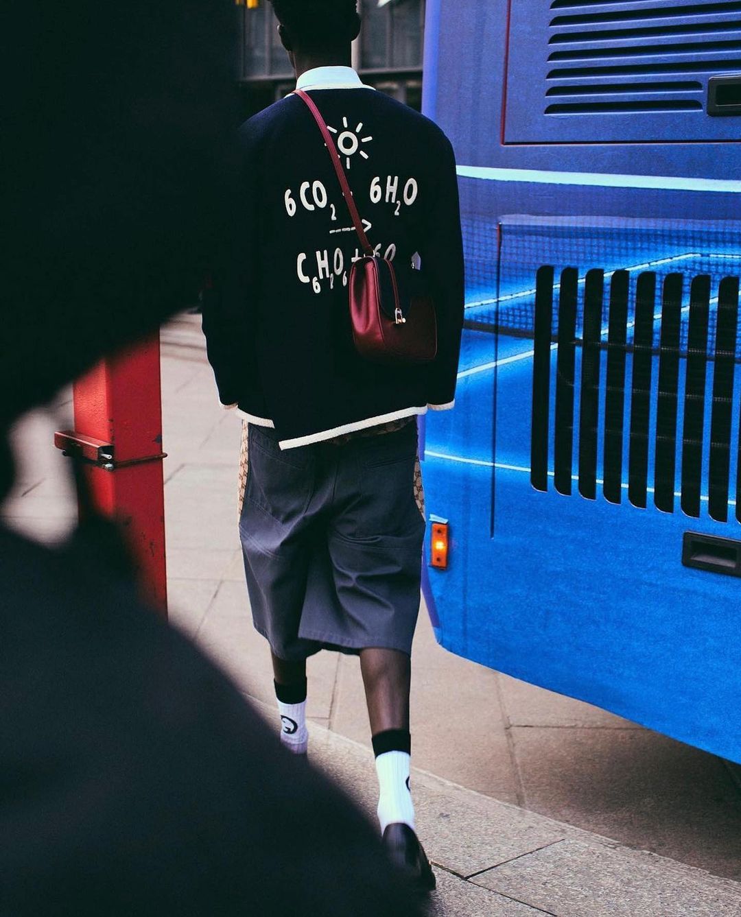 Person in streetwear wearing jacket with science equation walking past a blue bus on a city street, with red shoulder bag.