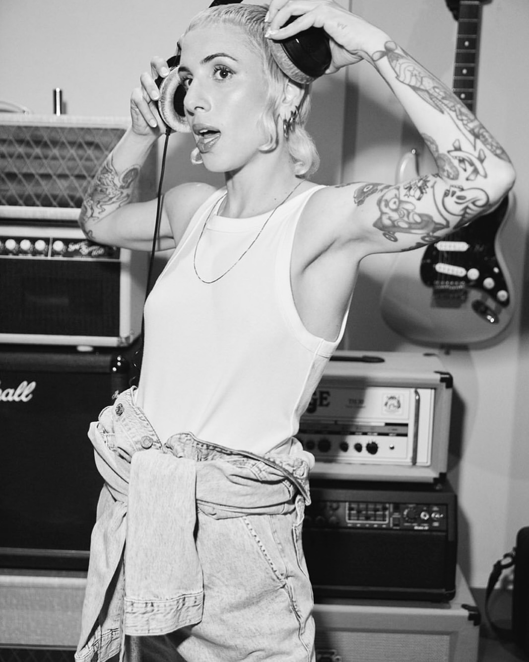Black and white portrait of a tattooed young woman with a tank top and overalls, wearing headphones, surrounded by music equipment indoors.