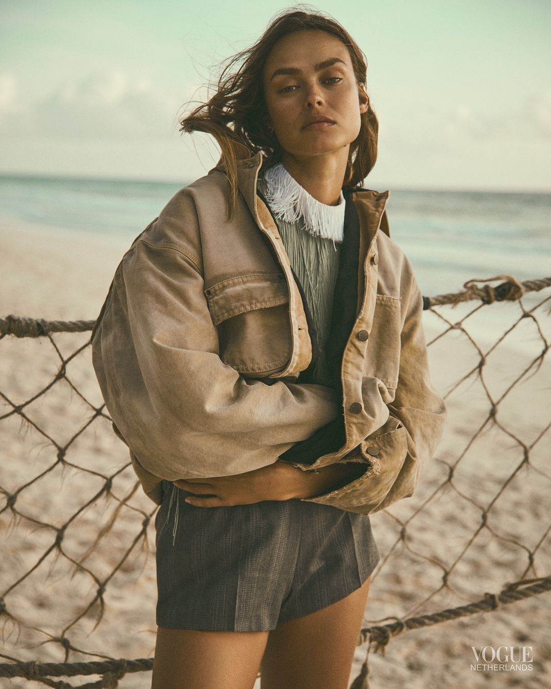 Model standing on a sandy beach wearing a beige jacket and grey shorts, photographed by Stefano Galuzzi.