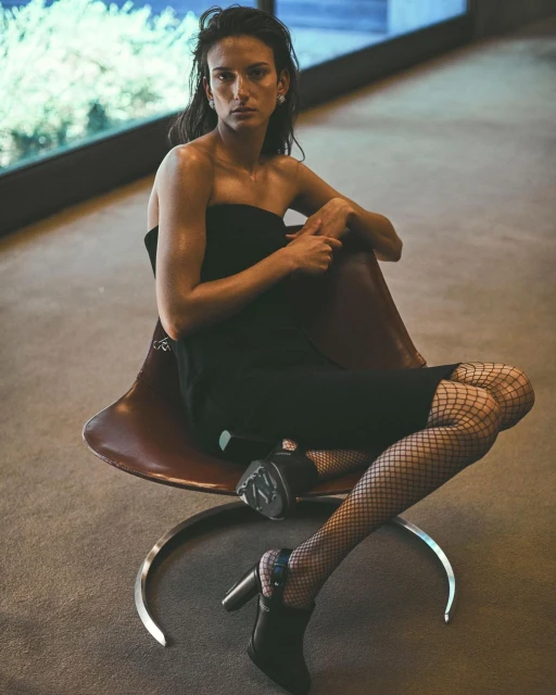 Indoor portrait of a solo adult woman in a strapless dress and fishnet stockings, sitting in a brown chair by a window.