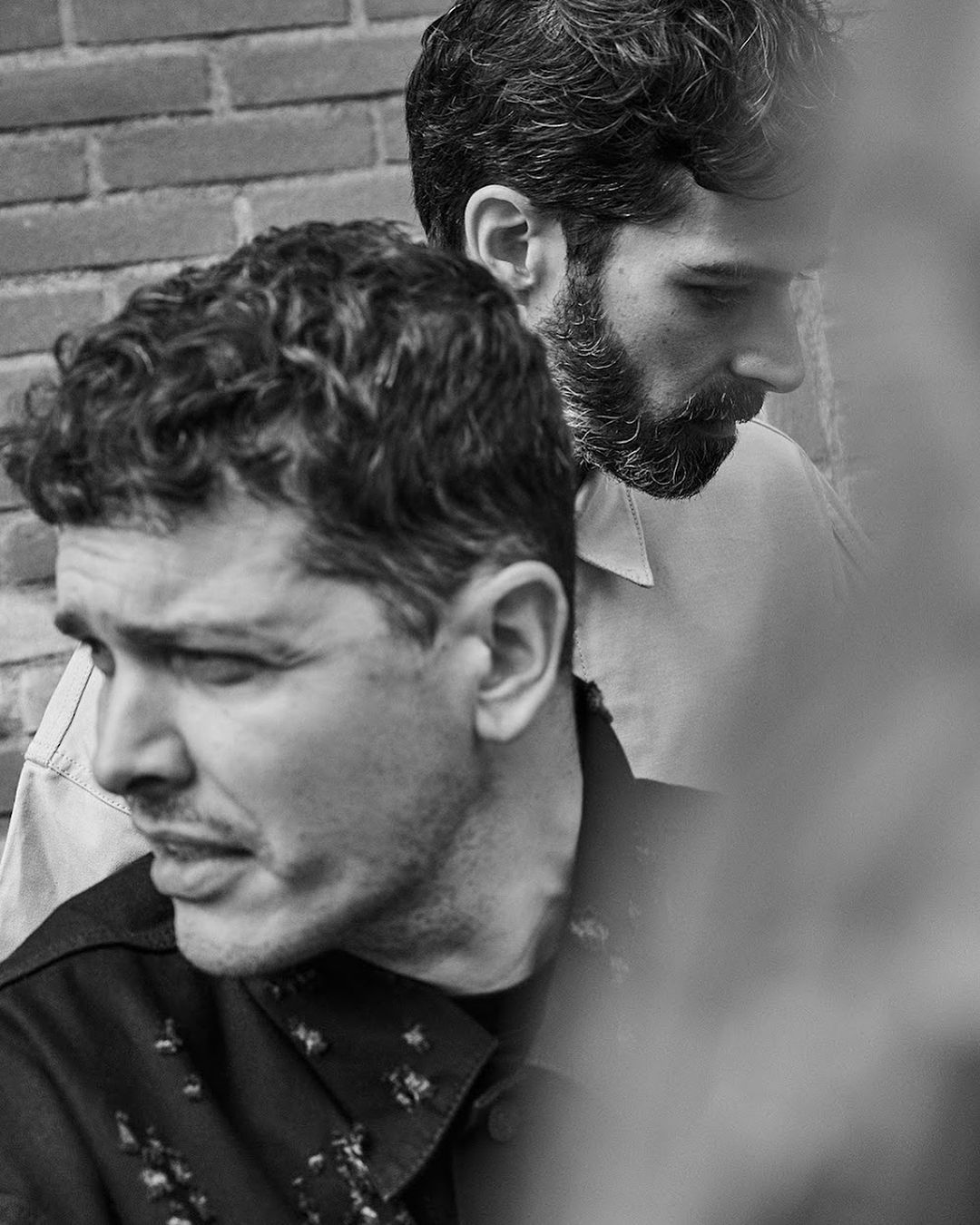 Black and white portrait of two men with short hair and beards posing outdoors near a brick wall.