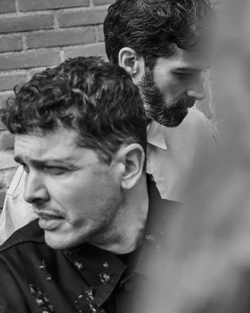Black and white portrait of two men with short hair and beards posing outdoors near a brick wall.