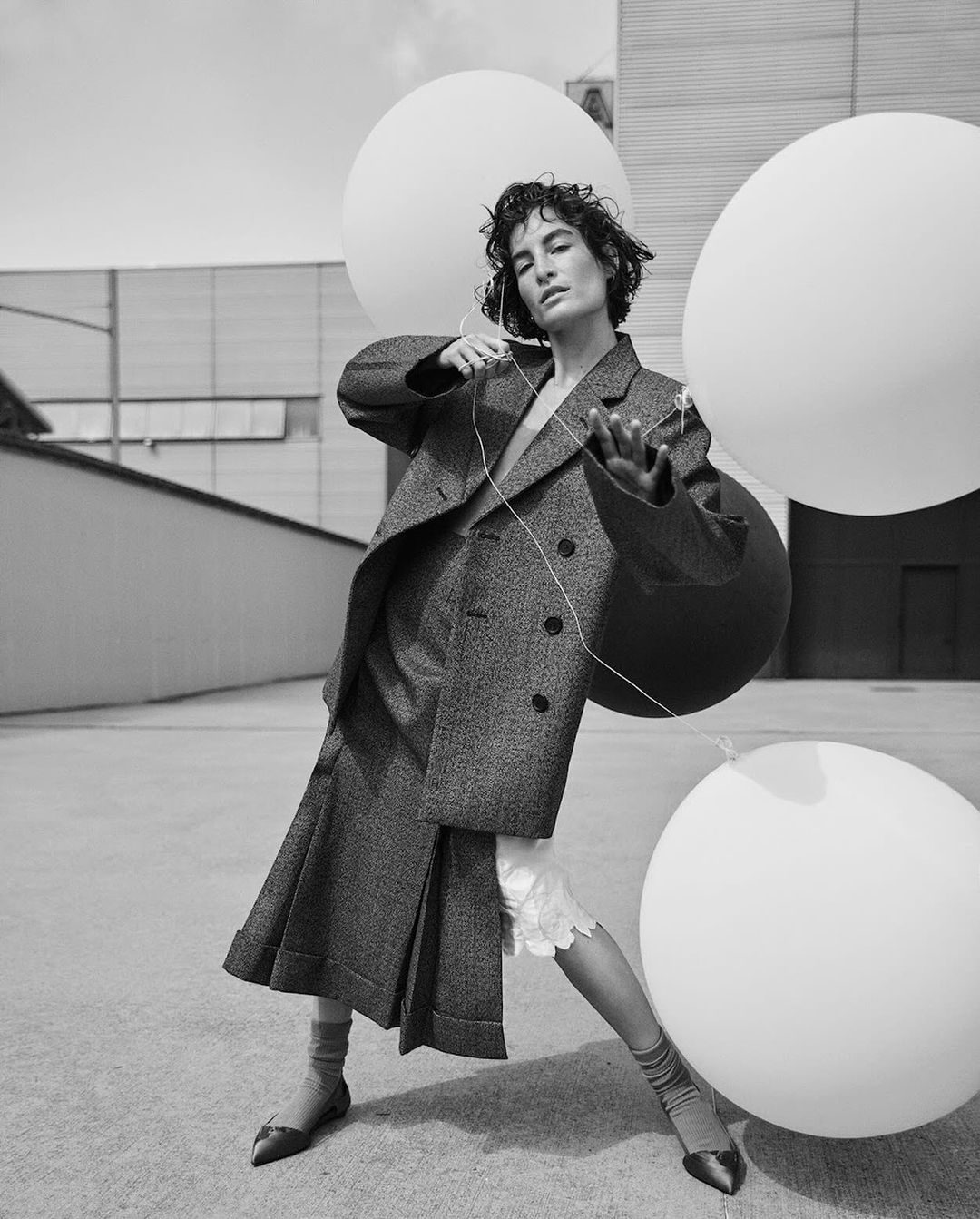 Black and white fashion shot of a female model outdoors wearing a tweed coat and skirt with balloons and connecting wired earphones.