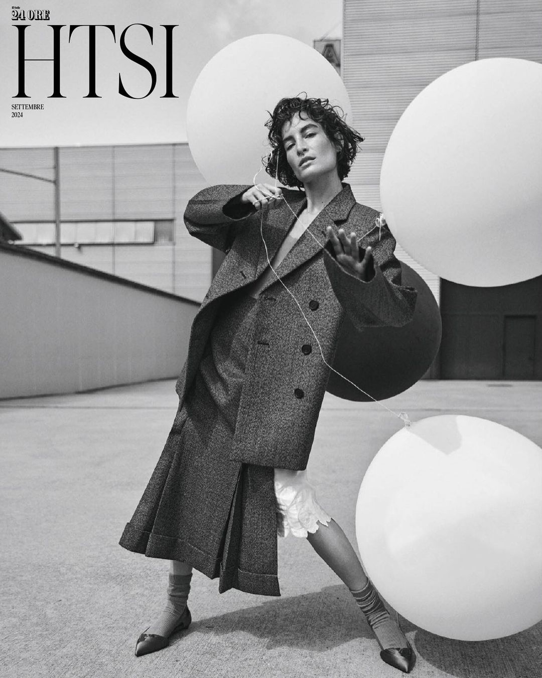 Model in oversized tweed blazer and pleated skirt holding large white balloons outdoors in a monochrome editorial image