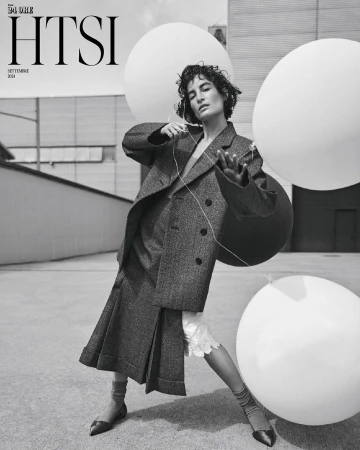 Model in oversized tweed blazer and pleated skirt holding large white balloons outdoors in a monochrome editorial image