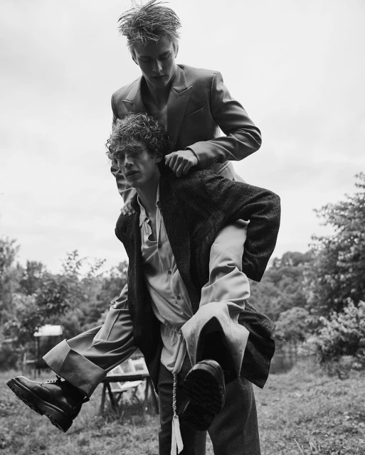 Two young adult men in an outdoor setting. One man gives the other a piggyback ride, both wearing stylish suits in black and white.