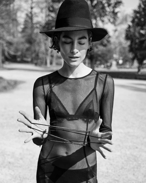Monochrome portrait of a person in a sheer dress and hat, holding a cat's cradle string game, captured in an outdoor park setting.