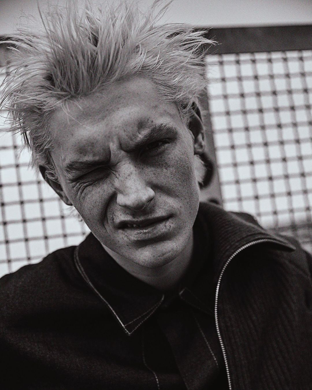 Black and white portrait of a young adult male with spiky blonde hair, fair skin, and a scowling facial expression in front of a mesh background.