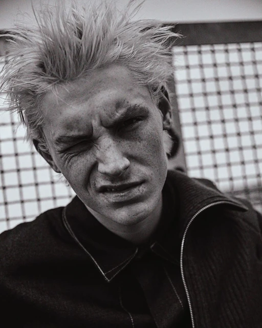 Black and white portrait of a young adult male with spiky blonde hair, fair skin, and a scowling facial expression in front of a mesh background.