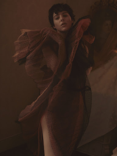 Fashion portrait of a woman with short hair in a sheer draped dress, posing in a warm, muted color palette with soft lighting.
