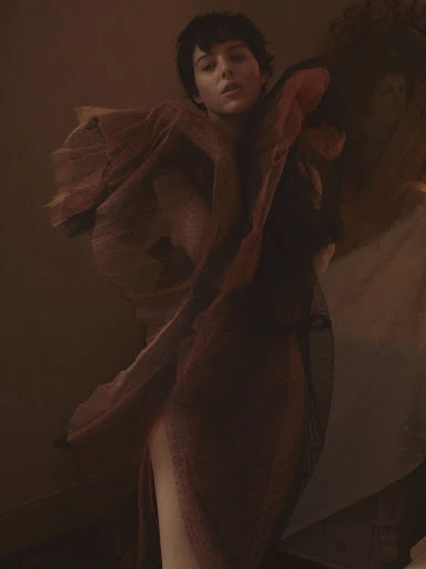 Fashion portrait of a woman with short hair in a sheer draped dress, posing in a warm, muted color palette with soft lighting.