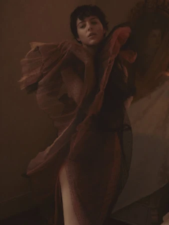 Fashion portrait of a woman with short hair in a sheer draped dress, posing in a warm, muted color palette with soft lighting.