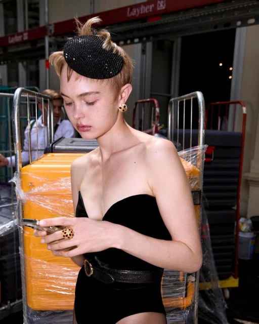 Young woman in a black strapless swimsuit and sparkly hat holding a phone indoors, surrounded by equipment and wearing ring jewelry.