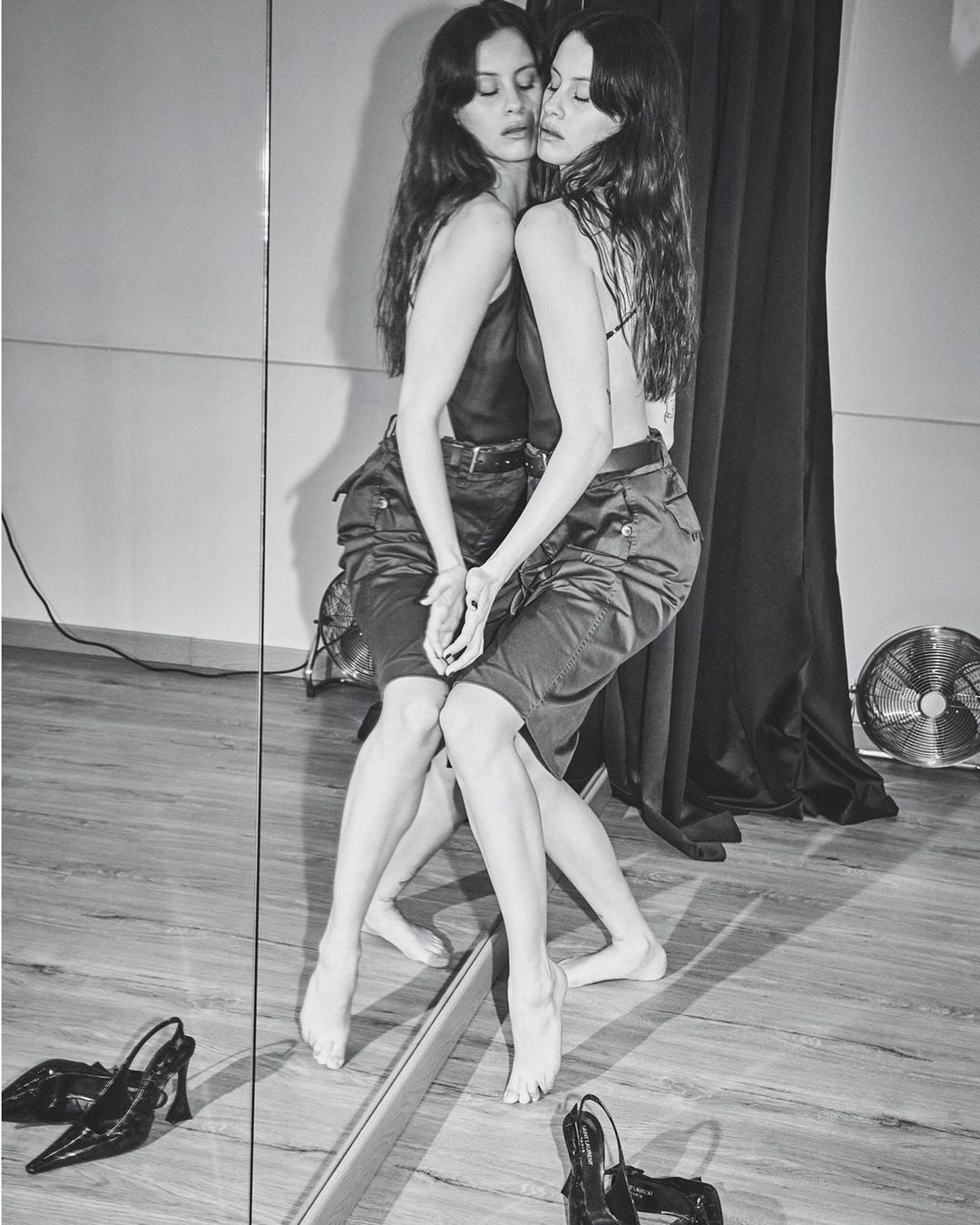 Black and white portrait of a woman reflected in a mirror wearing a dark top and cargo shorts with heels on the wooden floor.