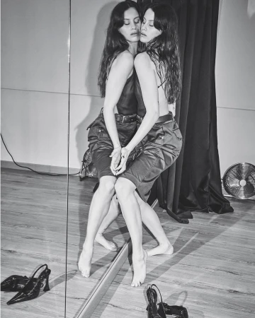 Black and white portrait of a woman reflected in a mirror wearing a dark top and cargo shorts with heels on the wooden floor.