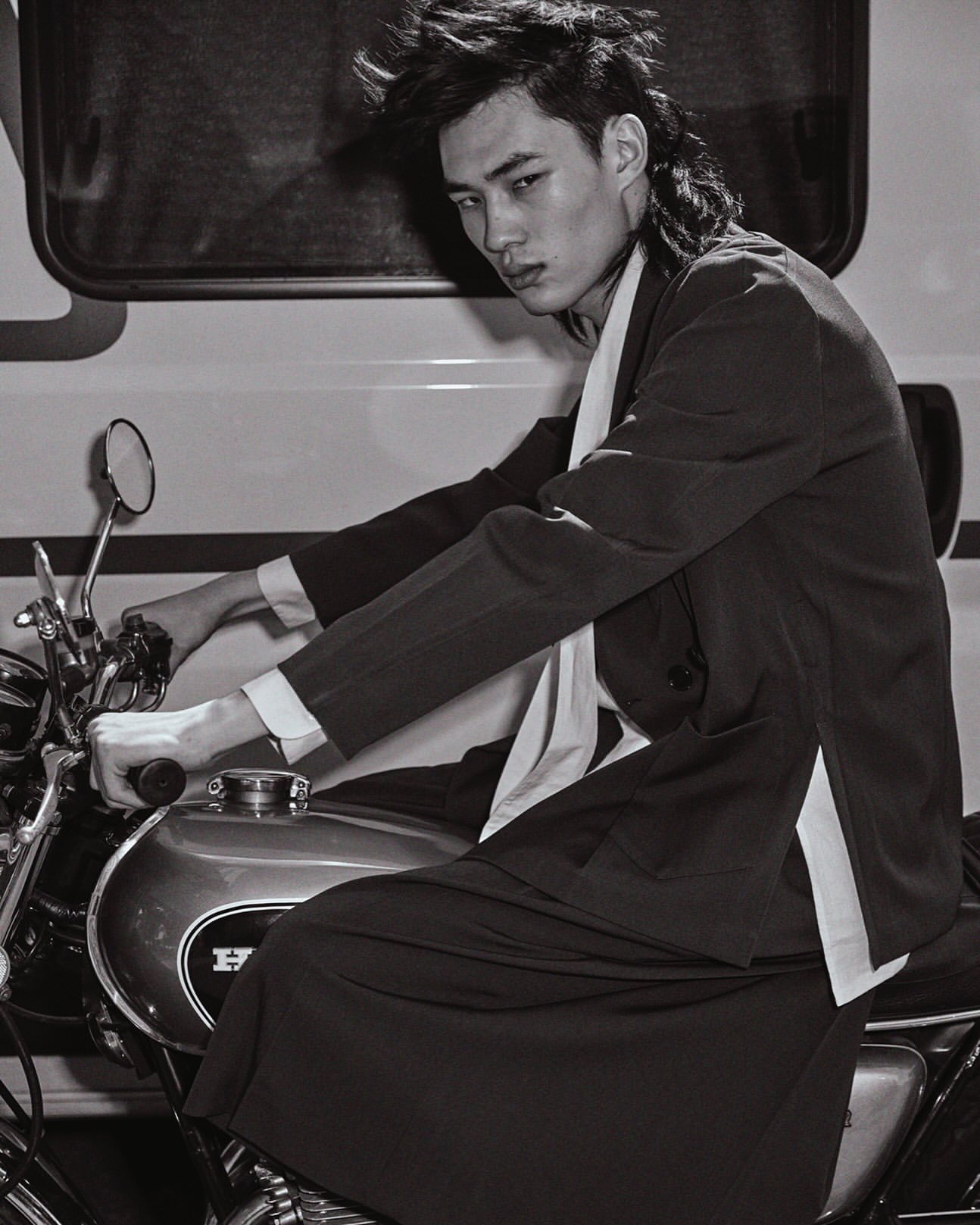 Monochrome shot of a young adult man in a suit sitting on a vintage Honda motorcycle, with a trailer visible in the background.